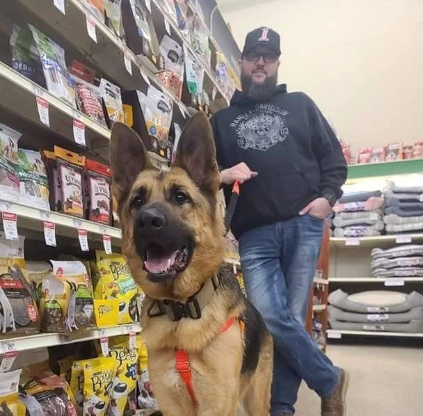An Operation Saves team member standing in a pet supply aisle with his happy German Shepherd wearing a red harness.